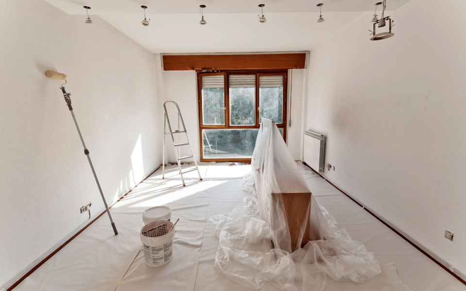 Habitación preparada para ser pintada, con suelos y muebles cubiertos con plástico protector, un rodillo de pintura y una escalera junto a la ventana que ilumina el espacio.